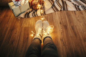 girl legs in stylish warm sock sitting with garland lights at christmas tree with gifts. socks on...