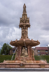 Fototapeta premium Glasgow, Scotland, UK - June 17, 2012: Doulton Fountain on Glasgow Green under heavy cloudscape. Green foliage and Pendleton building in back.
