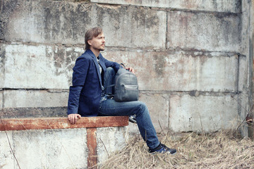 Handsome man in casual wear with modern leather backpack sits near concrete wall. Trendy accessory