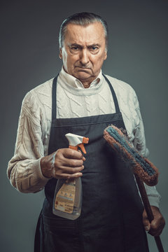 Senior Old Man Disgusted By The Idea Of Cleaning, Holding Cleaning Chemicals And Wearing Apron