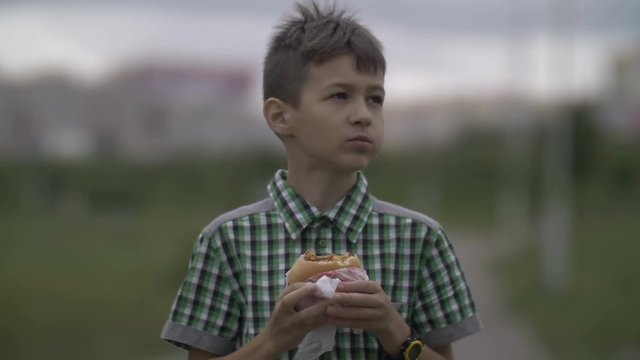 boy walks along the road in the street and eats a burger in the evening.