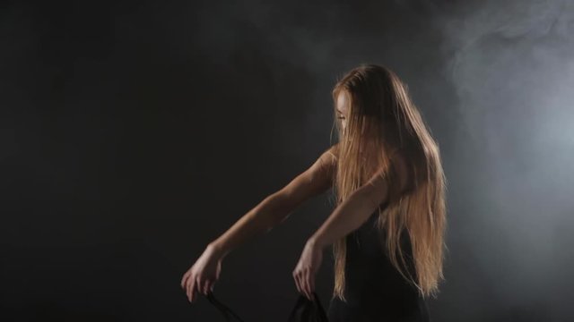 Professional ballerina dancing ballet in spotlights smoke on big stage. Beautiful caucasian young girl with long hair wearing black tight dress on floodlights background.