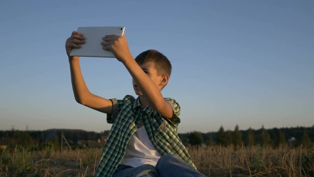 boy talking on video call on tablet sitting in field at sunset