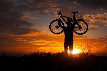 Obraz premium Silhouette the man stand in action lifting bicycle above his head on the meadow with sunset
