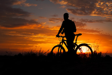 Obraz na płótnie Canvas Silhouette of sports person cycling on the meadow on the beautiful sunset. Young man with camera and bicycle.