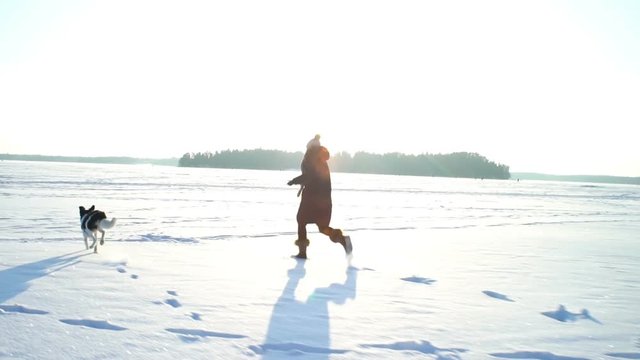 Winter fun snow vacation. Young woman with little dog running on the ice of the frozen sea, lake in sunny day in Scandinavia. Slow motion
