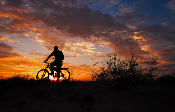 Silhouette of sports person cycling on the meadow on the beautiful sunset. Young man with camera and bicycle.