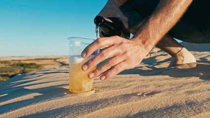 young man pouring beer from a bottle to a plastic cup glass outdoor in the desert sand dunes