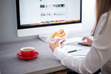 Young business woman eating burger with coffee at the workplace in office