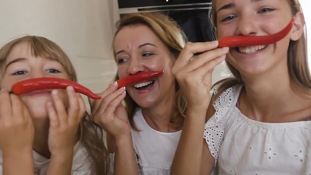 Two sisters and their mother dressed in white clothes are having fun with chili pepper at home in the kitchen. Girls make mustache from red pepper chili and have fun looking at the camera
