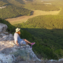 Fototapeta na wymiar A traveler girl with a hat on her head sits on top of a mountain. Active rest concept. Summer view of the mountains with green trees in sunlight