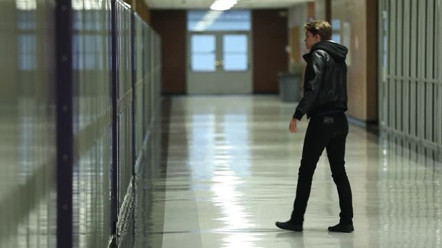 Teenager walks over to lockers and sits on the ground alone in empty school hallway.