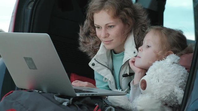 Beautiful young woman and her little daughter are sitting in the open trunk of a car on the river bank of the sea enjoying a laptop