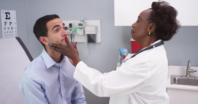Professional black MD checking young patients neck for swollen thyroid and lymph nodes. Young latino male patient having routine health checkup