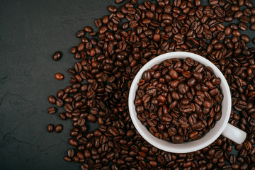 White cup full of with roasted brown coffee beans on the black concrete stone background. Flatlay...