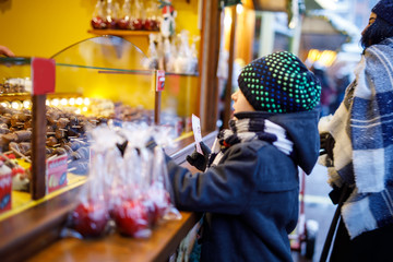 Little cute kid boy near sweet stand buying sugared apples and chocolate fruits. Happy child on Christmas market in Germany. Traditional leisure for families on xmas. Holiday, celebration, tradition.