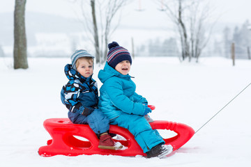 Two kid boys having fun sleigh ride during snowfall. Children sledding on snow. siblings riding a sledge. Twins play outdoors. Cute little toddlers sled on winter day. Active fun for family vacation