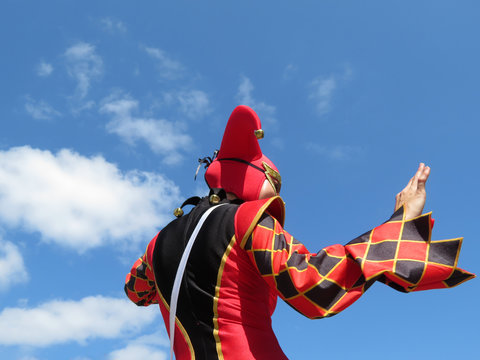 Person in a jester costume isolated on blue sky with white clouds, carnival concept. Harlequin in black-red foolscap with bells, girl dressed as a Joker