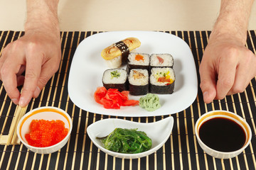 Hands with chopsticks and sushi, chuka salad, soy sauce and salmon caviar on a black bamboo mat.