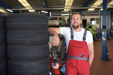 Reifenwechsel in der Autowerkstatt - Portrait fröhlicher  Mechaniker neben einem Reifenstapel im...