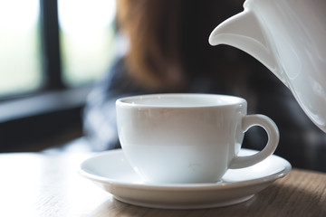 Closeup image of a person holding teapot and pouring tea into a white cup on wooden table