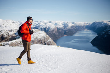 Traveling hiker man in red jacket walking on the snowy mountain top near Preikestolen, Pulpit Rock in Norway. Lysefjord and mountains. Active traveling concept