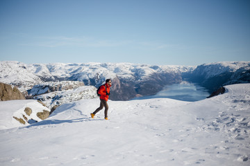 Traveler man in red jacket walking on the snowy mountain top near Preikestolen, Pulpit Rock in Norway. Lysefjord and mountains. Active traveling concept