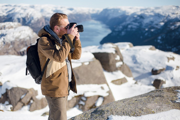 Traveler man taking photo on the camera on the top view of the Pulpit Rock, Preikestolen with beautiful landscape view of fjord between rocky shore with snow. Norwegian mountains. Lysefjord, Norway