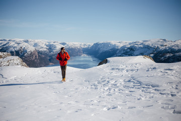 Young hiker man in red jacket walking on the snowy mountain top near Preikestolen, Pulpit Rock in Norway. Lysefjord and mountains. Active traveling concept