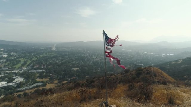 Aerial view circling a weathered American flag on a tall pole at the top of a dry golden hill. The frayed banner waves in a proud patriotic scene above a hazy California valley of houses.