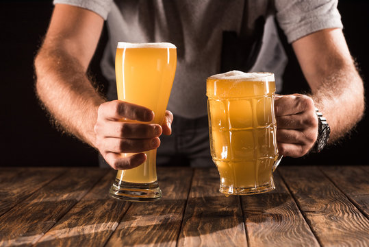 cropped image of man putting two mugs with fresh beer with foam at wooden table