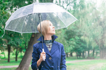 A woman of 30-40 years in a blue cloak goes under a transparent umbrella in the park.