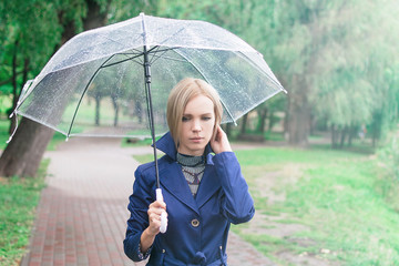 A woman of 30-40 years in a blue cloak goes under a transparent umbrella in the park.