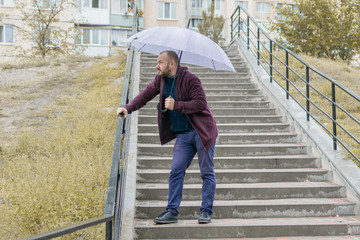 An unshaven man of 35-40 years old in a sweatsuit descends the stairs under an umbrella.