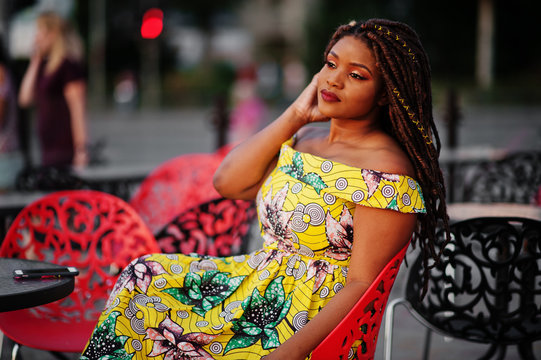 Cute small height african american girl with dreadlocks, wear at coloured yellow dress, sitting at outdoor cafe on red chair.