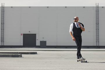 Confident young businessman in business suit on longboard hurrying to his office, on the street in...