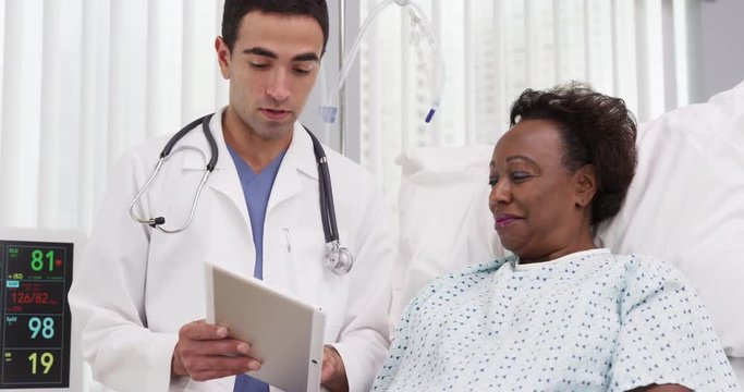 Positive female black patient recovering in hospital bed looking at tablet. Charming young latino doctor using electronic notebook tablet to share patients surgery results