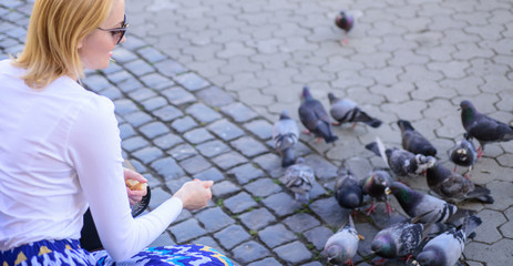 Group doves on city square waiting treats. Girl feeding dove birds urban background. Girl blonde...