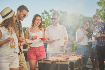 Group of people standing around grill, chatting, drinking and eating. 