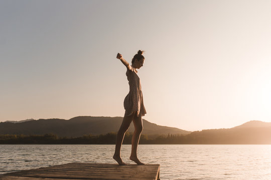 Side view of young caucasian girl dancing on the edge of a dock of a lake in a sunset of a day of summer mountains background