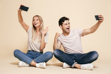 Young loving couple sitting isolated over beige wall background make selfie by mobile phones.