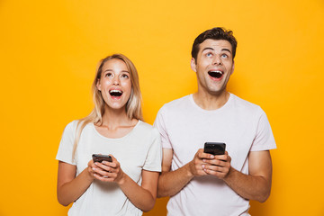 Happy excited young loving couple standing isolated over yellow wall background using mobile phones.