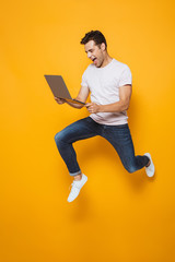 Young man jumping isolated over yellow wall background using laptop computer.