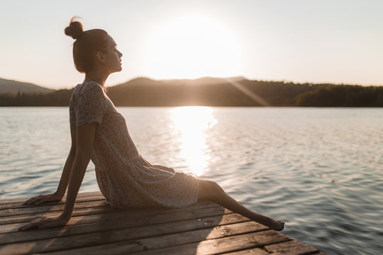 Young caucasian girl sitting and relaxing on a dock of a lake in a sunset of summer mountains background