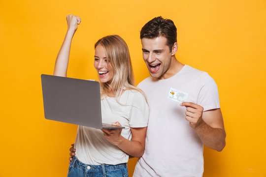 Young loving couple isolated over yellow wall background using laptop computer make winner gesture holding credit card.