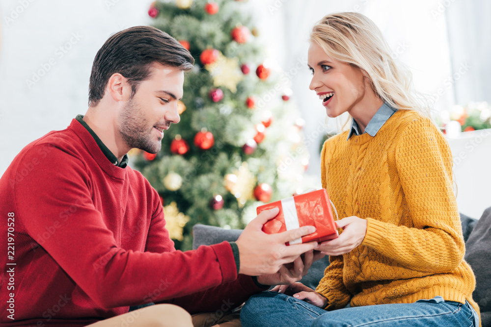 Wall mural man gifting present surprised to girlfriend at home with christmas tree