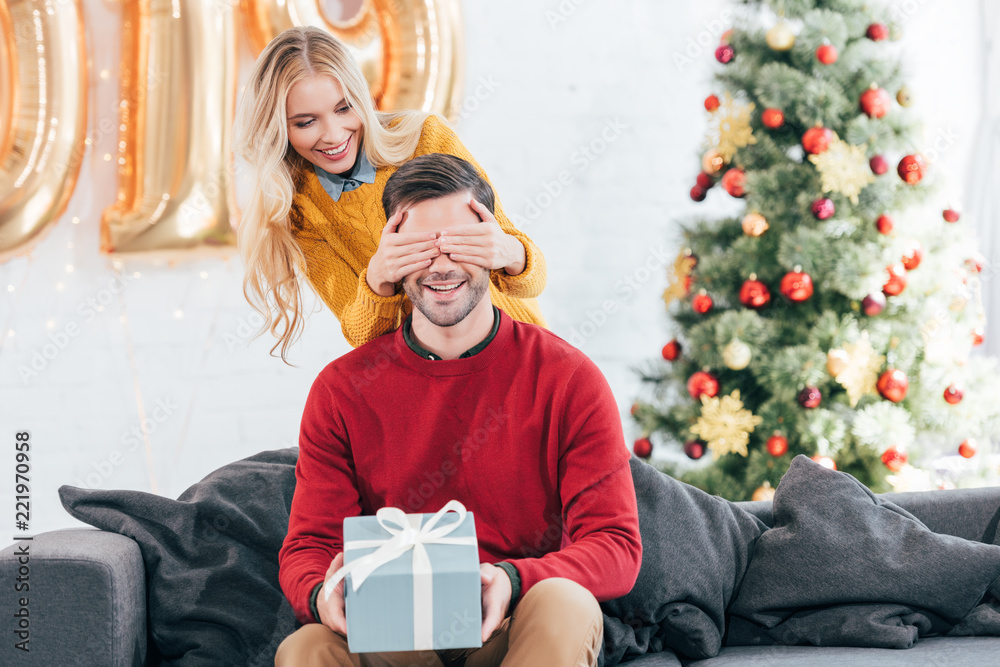 Wall mural girl closing eyes and making surprise with gift box for boyfriend at home with christmas tree