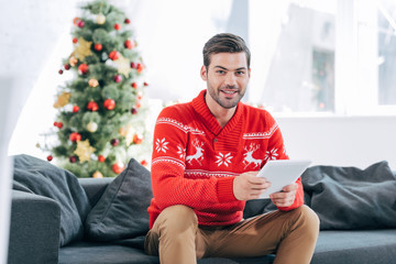 happy man using digital tablet and sitting on sofa with christmas tree behind