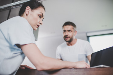 Fototapeta premium low angle view of girlfriend using laptop, irritated boyfriend looking at her in kitchen
