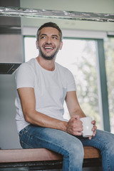 Fototapeta premium smiling handsome man sitting on kitchen counter with cup of tea in morning and looking away